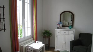 A room with a white table and chairs, a green ottoman, a dark armchair, a white cabinet with a mirror, and a window with colorful curtains. - NORMAN HOUSE IN JUNO BEACH  (Bernières-Sur-Mer)