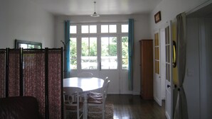 A dining area with a table and chairs, a window with curtains, and a door with a coat rack. - NORMAN HOUSE IN JUNO BEACH  (Bernières-Sur-Mer)