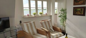 A living room with a sofa, a glass coffee table, a television, and a window with a view of a lake. - Apartment upper floor with balcony - Haus am Schlei Noor (Maasholm)