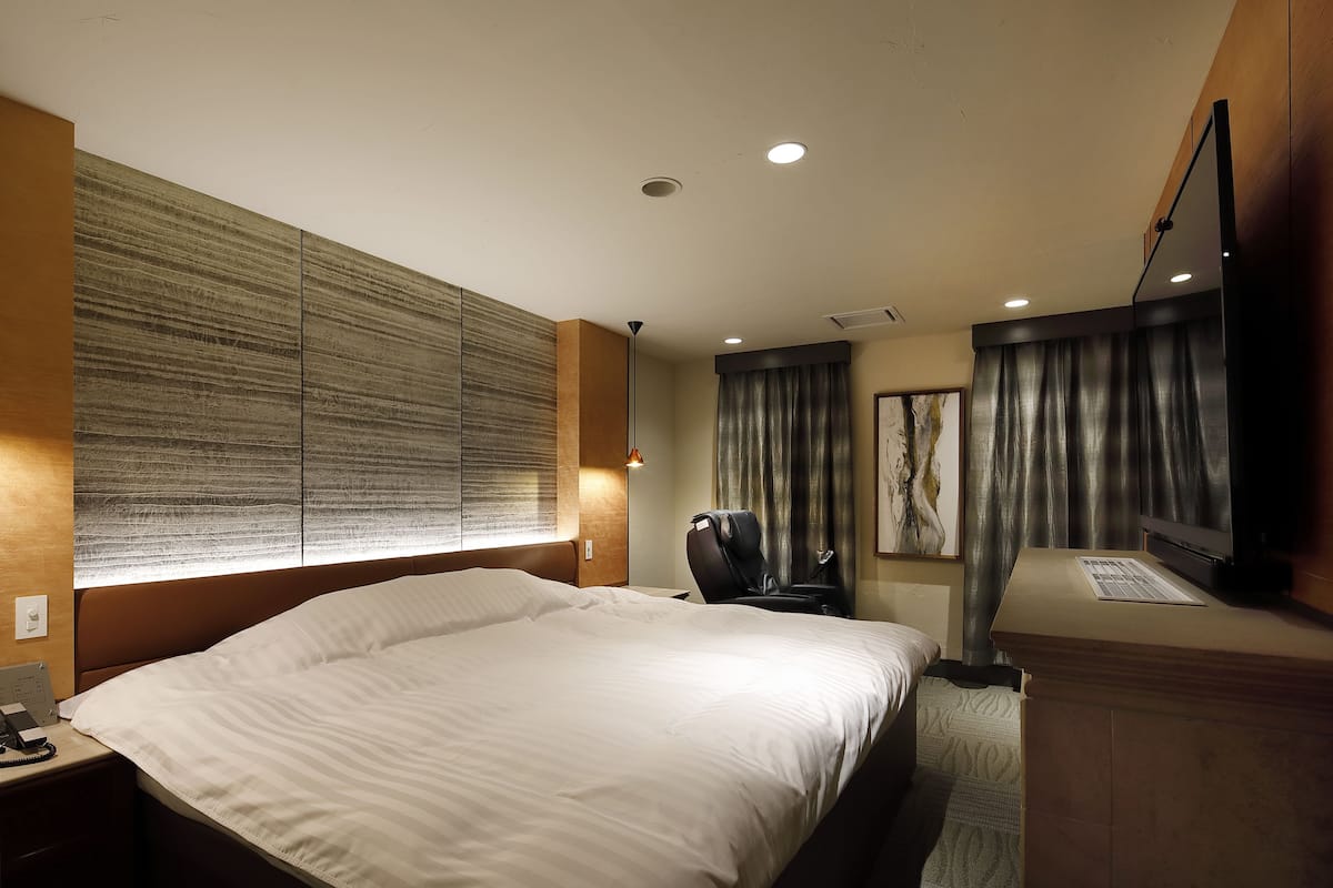A hotel room with a large bed, a desk with a flat-screen TV, a massage chair, and a large window with curtains.