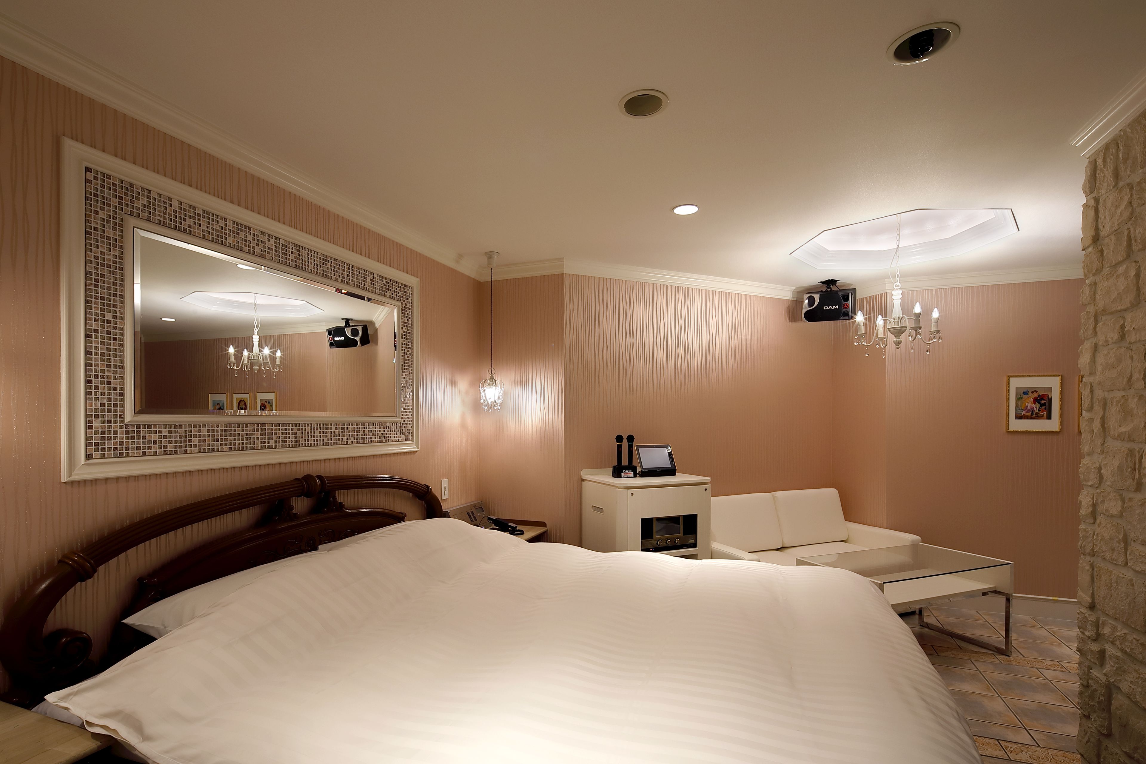A hotel room with a large bed, a sitting area with a chair and a small table, a mirror with a decorative frame, a chandelier, and a small cabinet with a television.