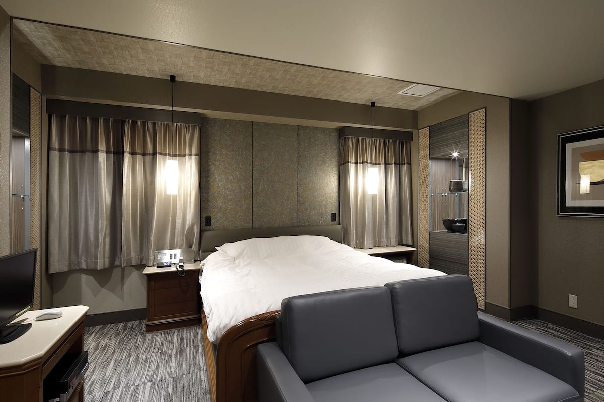 A hotel room with a bed, a desk with a computer, a chair, and a large mirror.