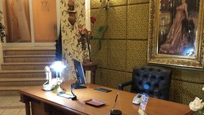 A reception desk with a computer, phone, and a vase of flowers. There is a framed painting on the wall and a staircase in the background.