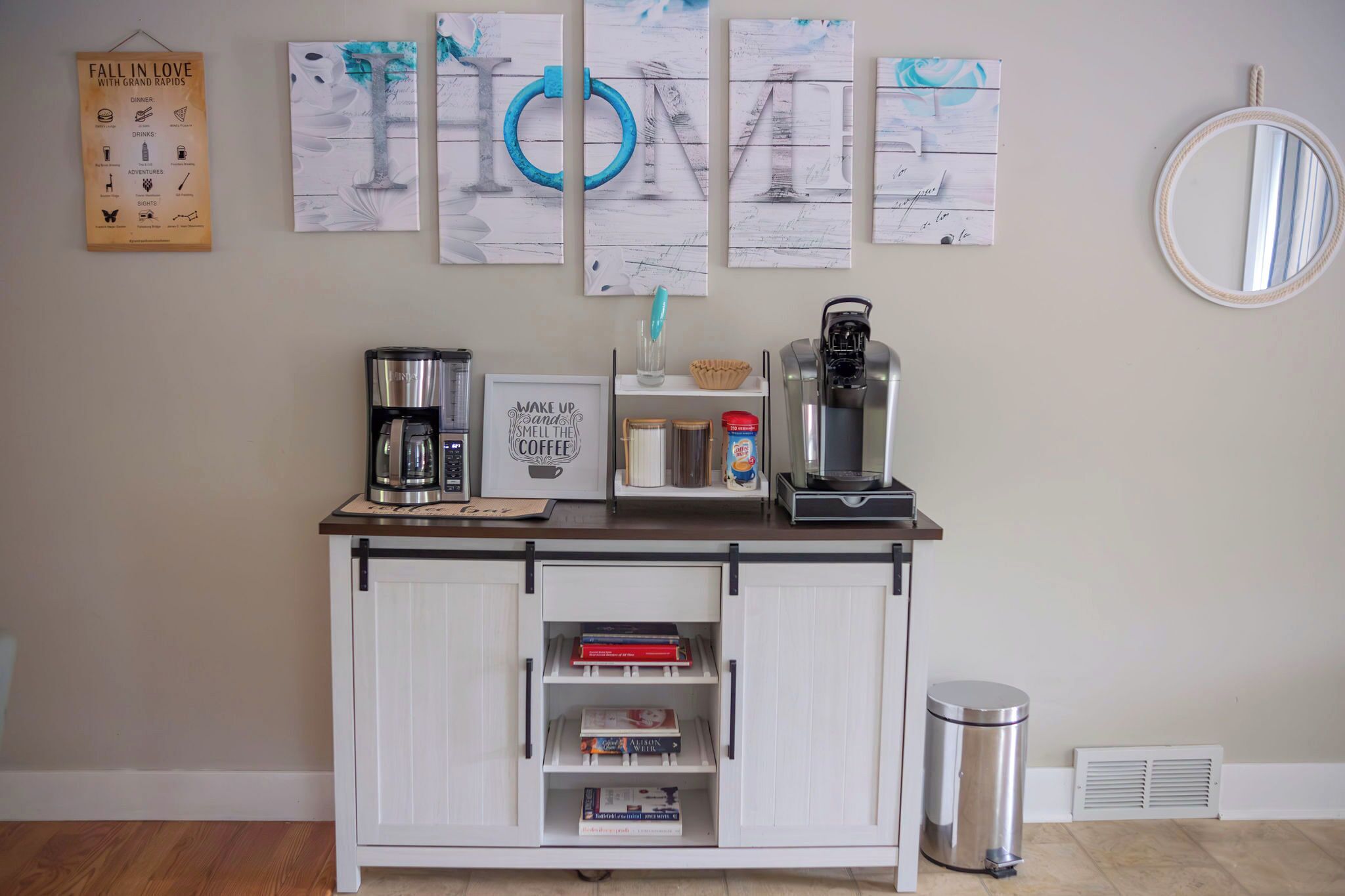 Una encimera de cocina con una cafetera, una jarra de café y una cita enmarcada. Debajo de la encimera, un armario blanco con libros.
