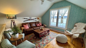 Una sala de estar estilo loft acogedora, con un sofá de cuero marrón, sillas con estampado, una mesa de centro de madera y un amplio ventanal con vistas a árboles.