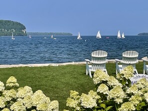 Une vue sur le bord de l’eau, avec plusieurs voiliers sur l’eau, une zone herbeuse avec des fauteuils Adirondack, et en arrière-plan, une île boisée.