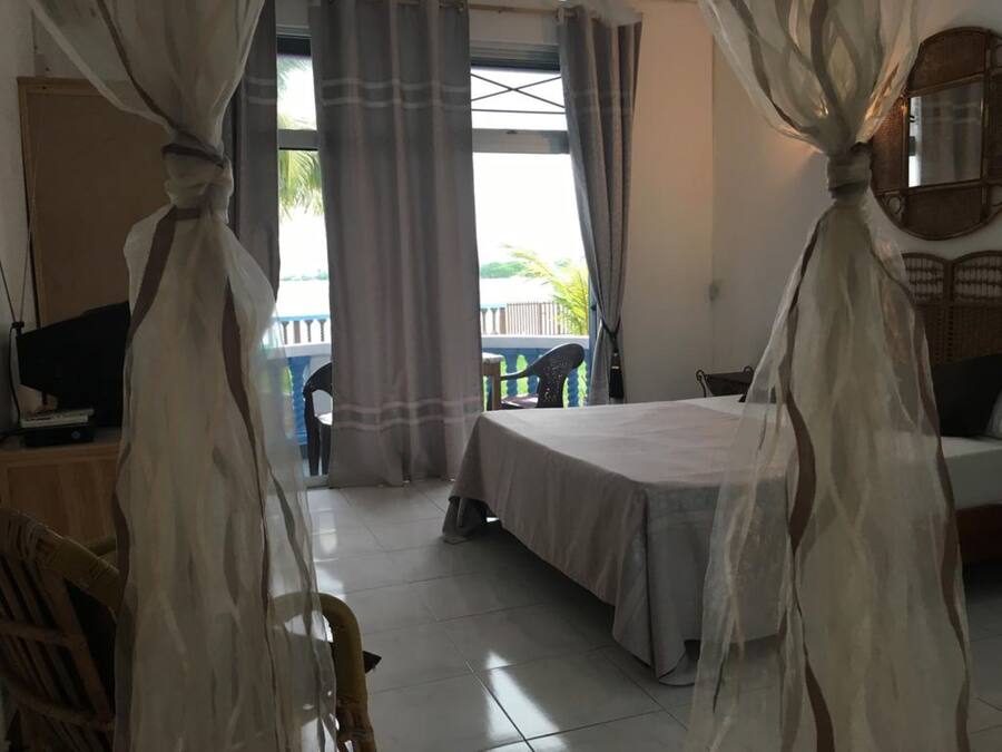 Un dormitorio con cama, escritorio con computadora y una silla. Tiene un balcón con barandilla y vistas a zonas verdes.