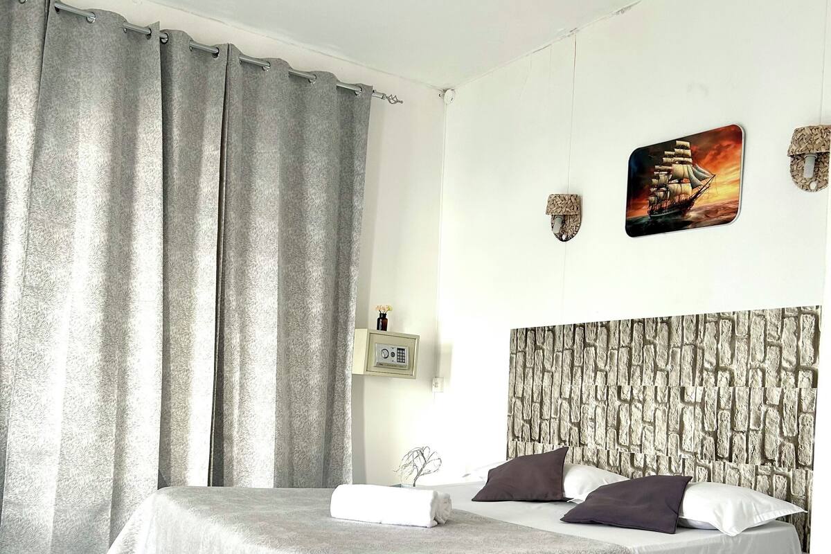 A neatly arranged bedroom with a bed, curtains, a ceiling fan, a wall-mounted clock, and a decorative ship painting.