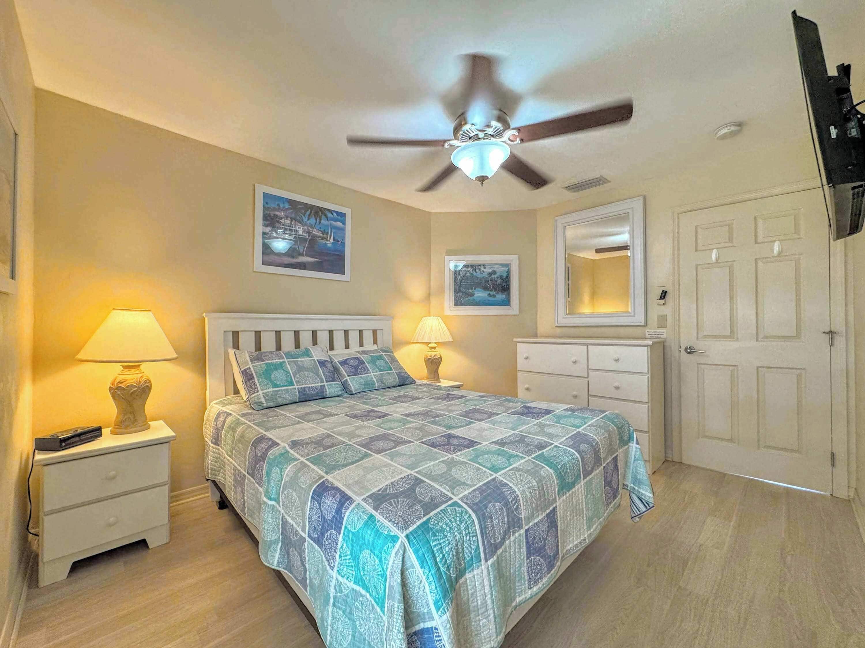 Une chambre à coucher comprenant un lit, des tables de chevet, un ventilateur de plafond, une commode et une porte.