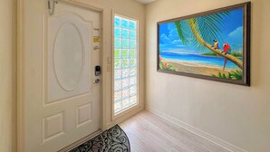 Un pasillo con una puerta blanca, un ventanal con vista a la playa y un cuadro con loros en un árbol.