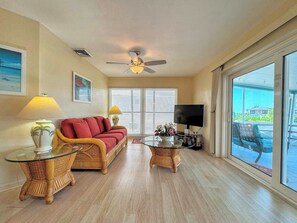 A living room with a sofa, two side tables, a ceiling fan, a television, and a large window. - 390 Bayland Canalfront Pool Home by Beach (Fort Myers Beach)