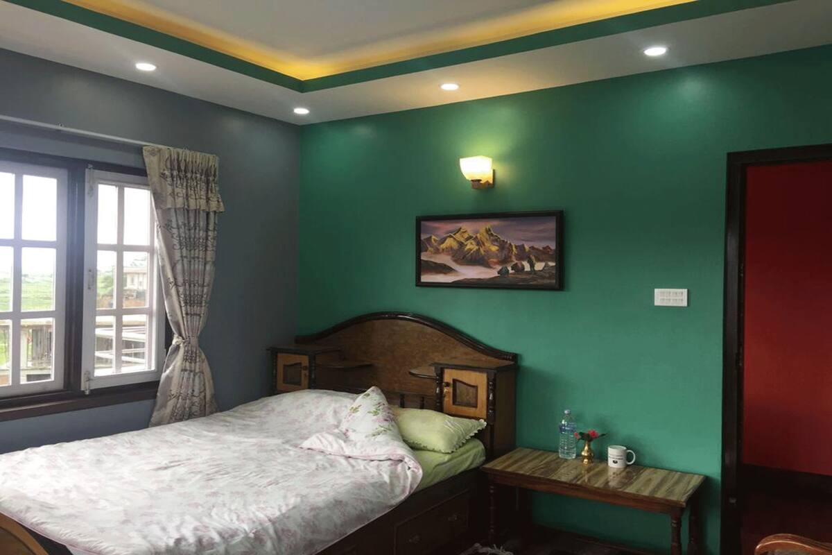 Une chambre à coucher avec un lit, une table de chevet, une lampe fixée au mur, un tableau et une fenêtre avec des rideaux.