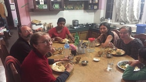 A round dining table with multiple people seated around it, plates of food, and various utensils. - Homestay Nepal (Kirtipur)