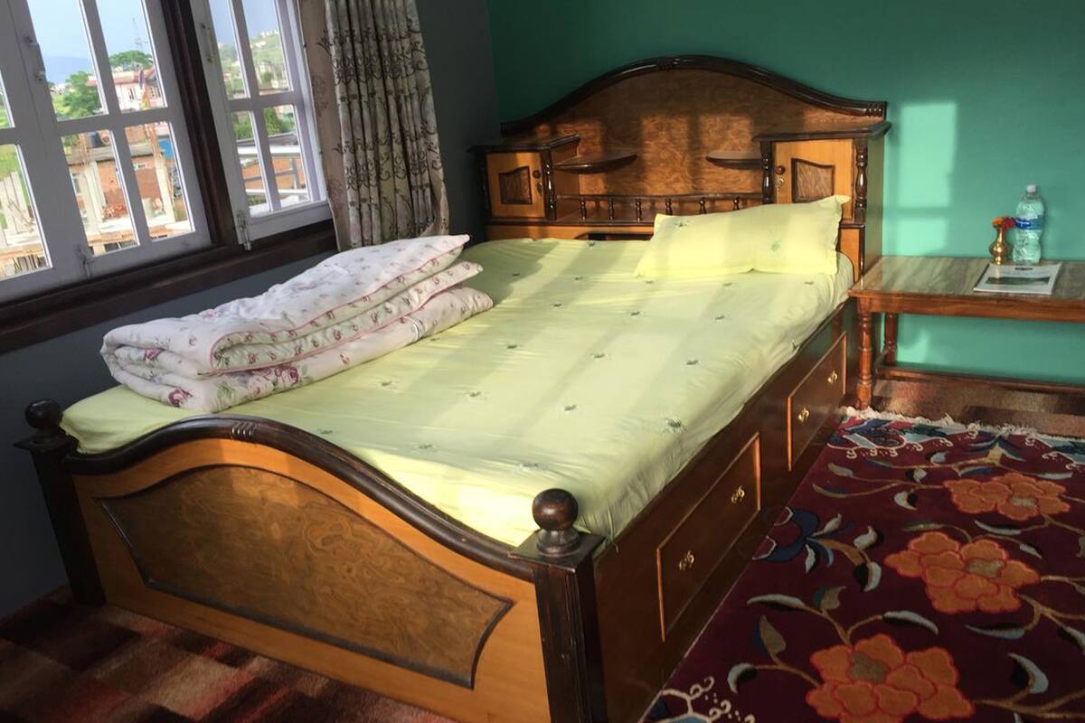 Un lit à baldaquin en bois, agrémenté d’une literie à motifs floraux, une table de chevet en bois avec une bouteille d’eau, et une vue sur l’extérieur par une fenêtre ornée de rideaux.
