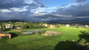 A rural landscape with green fields, scattered buildings, and a cloudy sky. - Homestay Nepal (Kirtipur)