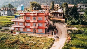A multi-story building with a red facade, surrounded by green fields and a hilly landscape. - Homestay Nepal (Kirtipur)