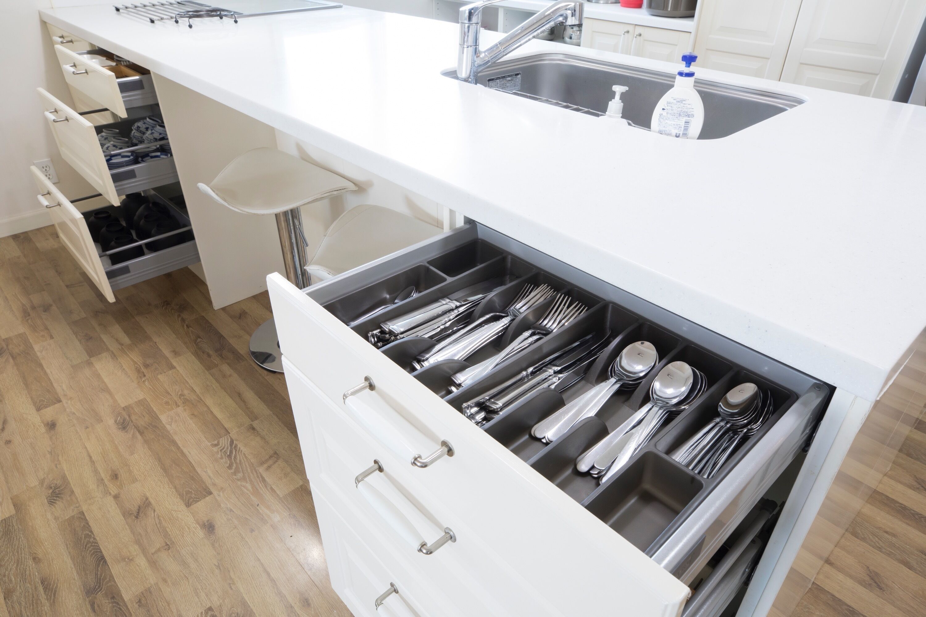 Una cocina con encimera blanca, pileta de acero inoxidable y un organizador de cajones con utensilios.