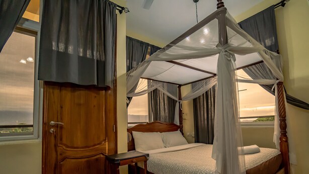 Una cama con dosel, una mesita de noche de madera y vista al mar por la ventana.