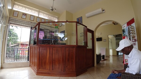 A reception desk with a wooden finish, a glass enclosure, and a sign indicating free WiFi. There is a clock on the wall and an air conditioning unit mounted above. - East Point Hotel Moshi (Moshi)