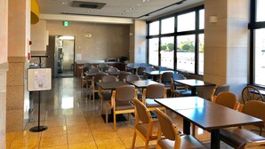 A spacious indoor dining area with multiple tables and chairs, a visible exit sign, and a large window providing a view of the outside. - Business Hotel Cosmo Inn (Fukuroi)
