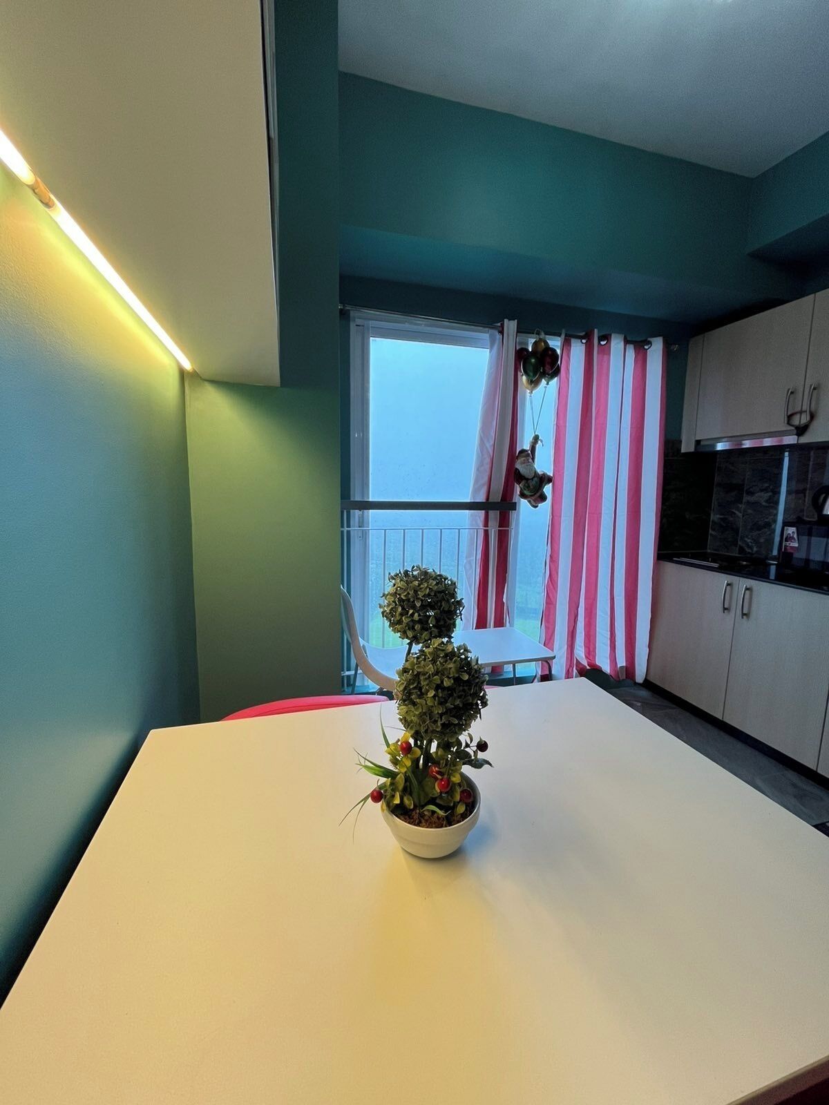 A modern kitchen with a white countertop, a potted plant, and a view of the outside through a window with striped curtains.