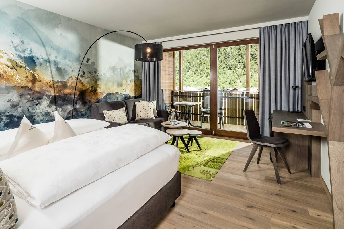 A modern hotel room with a large bed, a desk, and a balcony with a view of the outdoors.