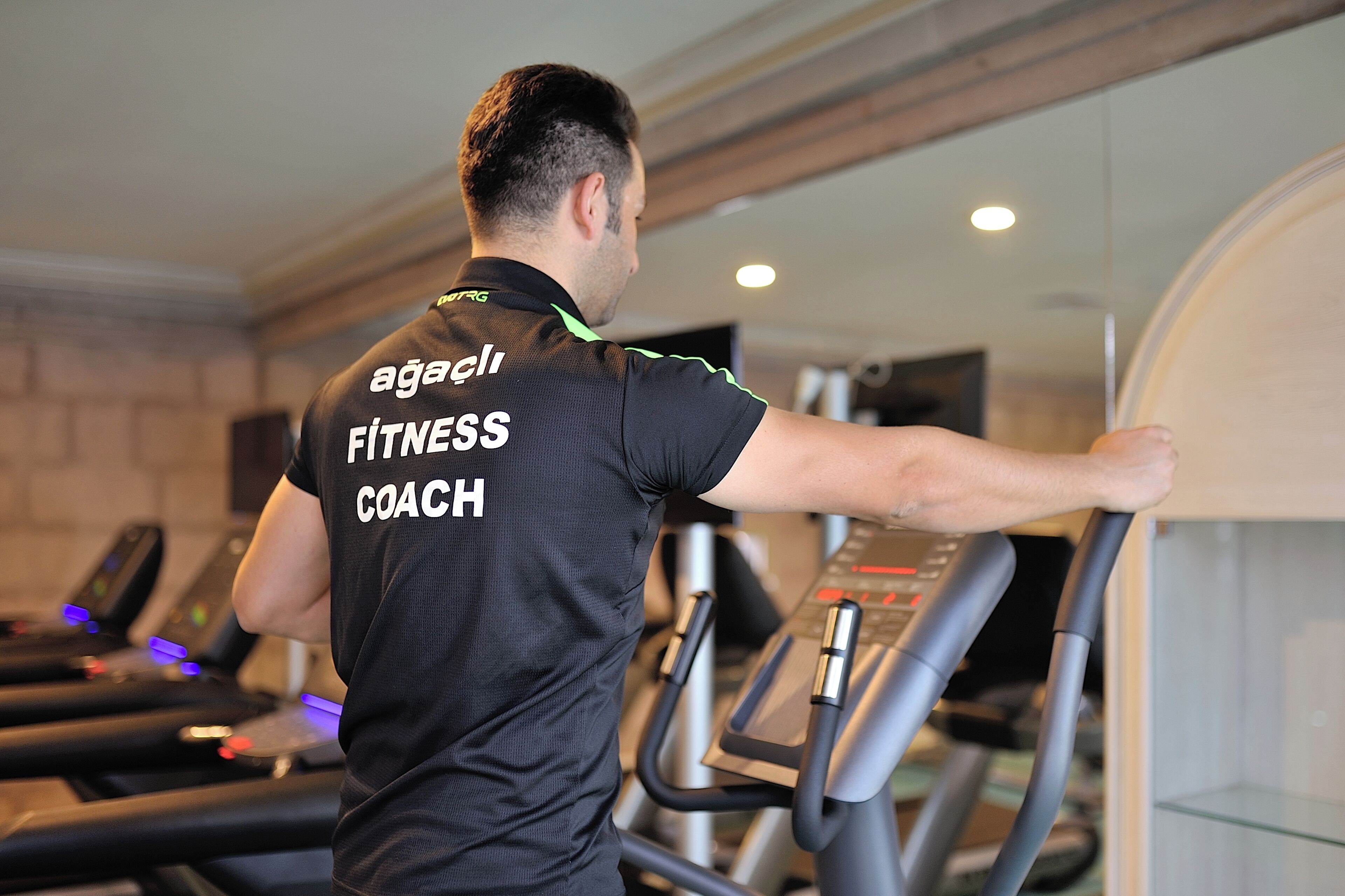 A fitness coach on a treadmill in a gym with exercise equipment and a brick wall.