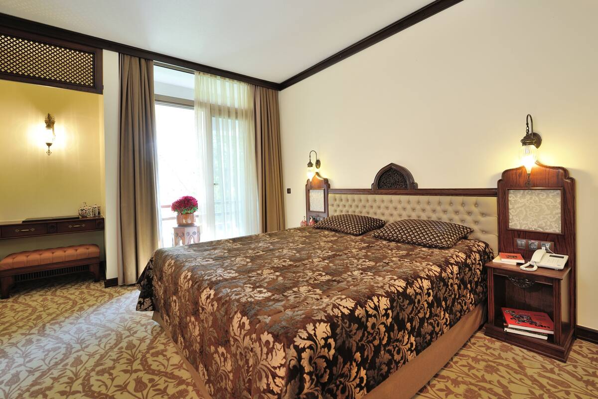 Una habitación de hotel con una cama grande, una mesita de noche con lámpara, un pequeño escritorio con teléfono y un banco.