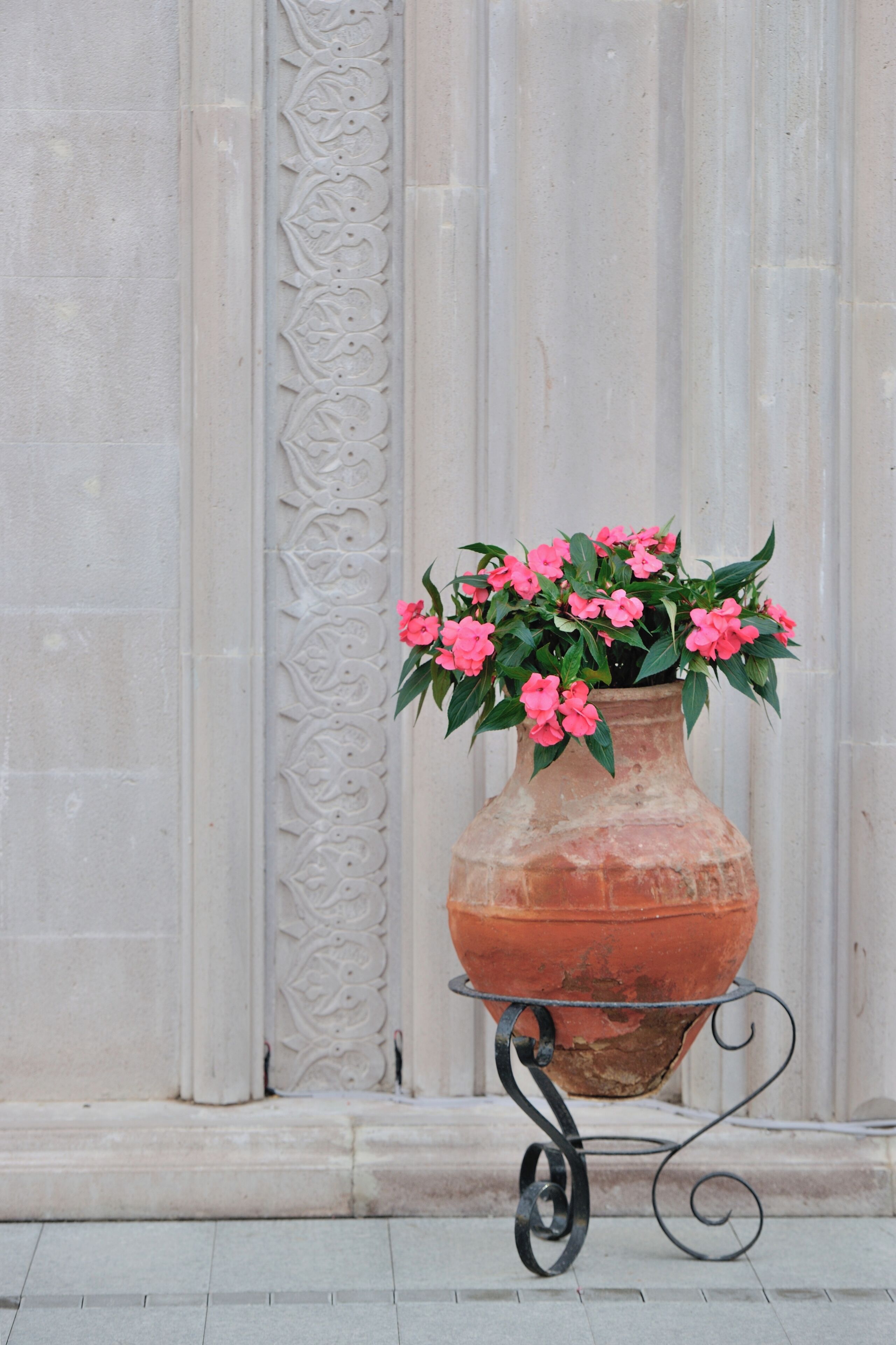 A terracotta pot with pink flowers on a metal stand against a white column with decorative carvings.