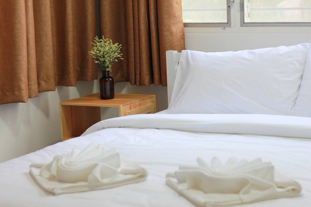 Una cama bien hecha con sábanas blancas, una mesita de noche de madera con un florero y cortinas marrones.