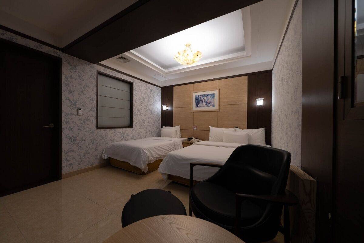 A hotel room with two beds, a desk, a chair, and a chandelier.
