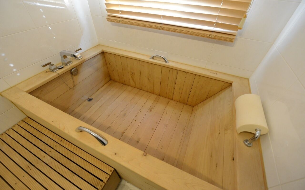 A wooden bathtub with a wooden bench, a toilet, and a towel dispenser.