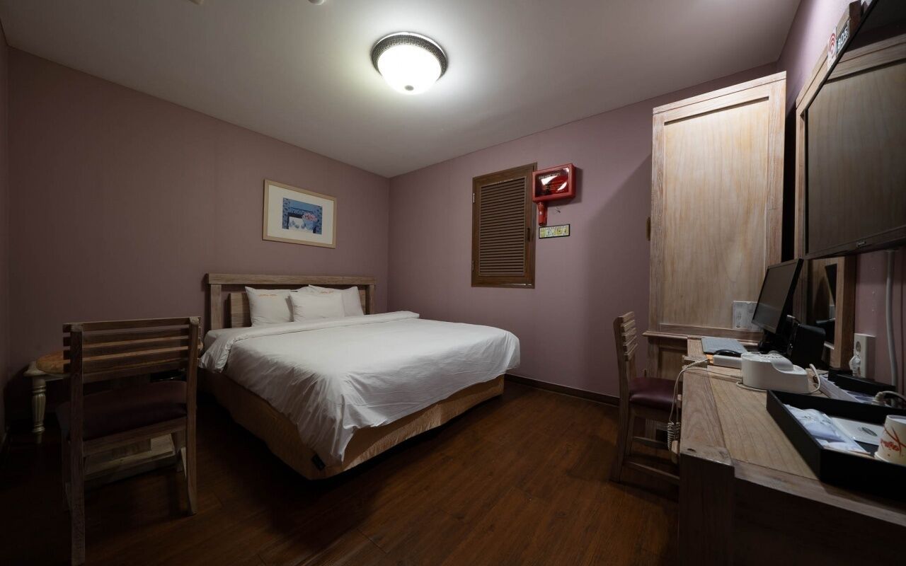 A bedroom with a bed, a desk with a computer, a chair, and a window with blinds.
