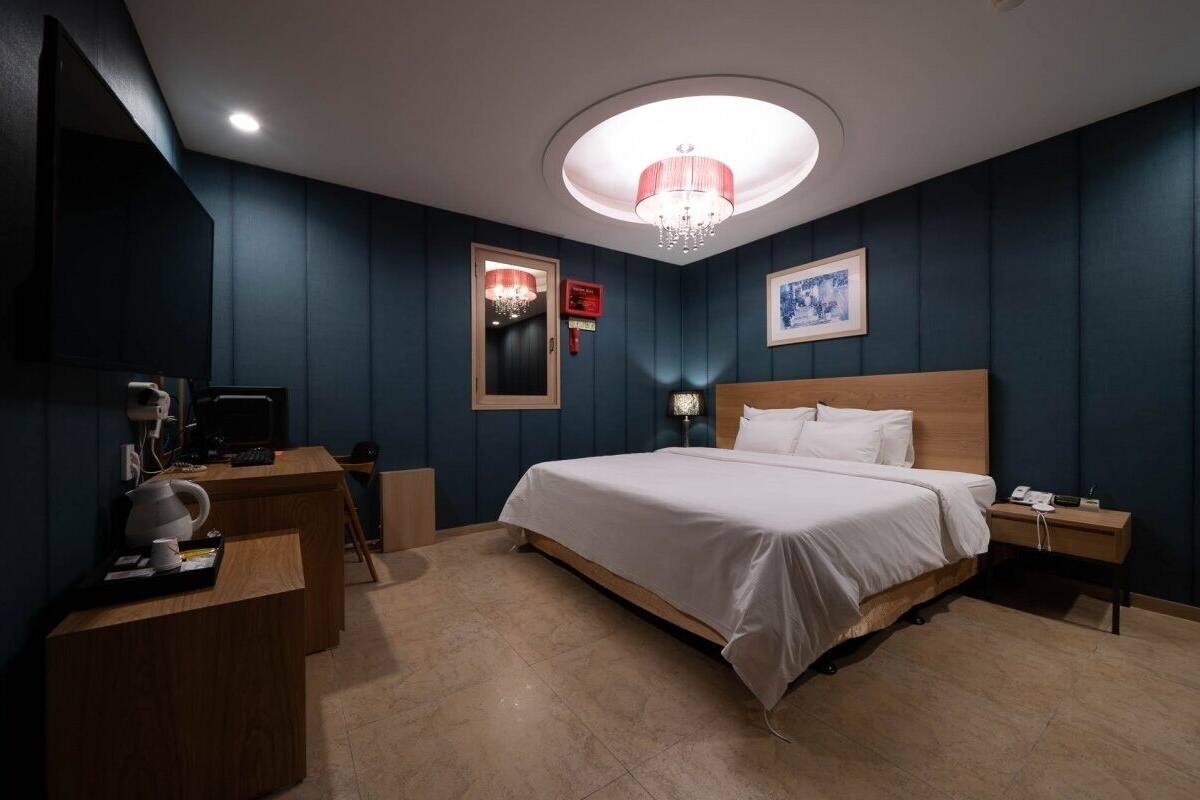 A hotel room with a large bed, a desk, a chair, a small table, a nightstand, a lamp, a chandelier, a framed picture, and a mirror.