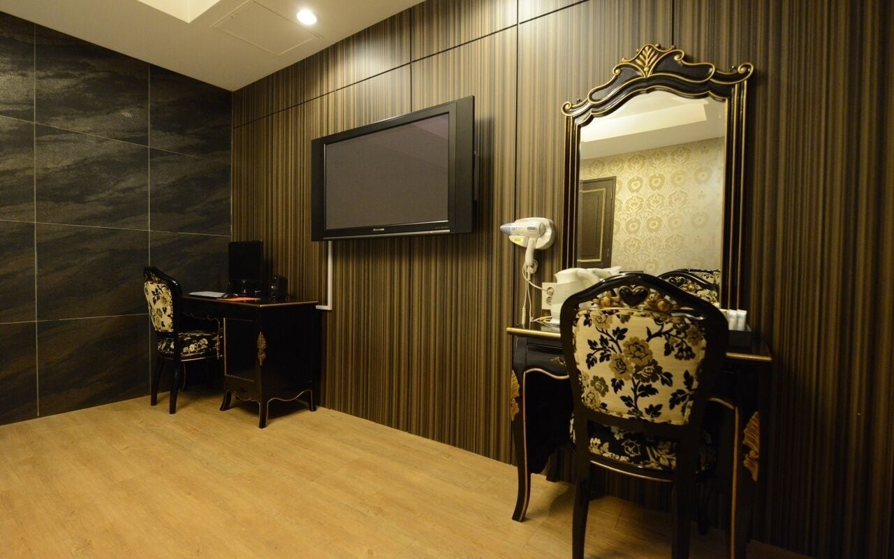 A hotel room with a large flat-screen TV mounted on the wall, a desk with a chair, a vanity with a mirror, and a bed with a headboard.