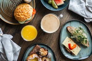 Une table dressée pour le petit-déjeuner, avec une variété d’aliments présentés sur des assiettes, notamment un sandwich, des toasts au fromage et un poivron rouge, ainsi qu’une assiette de viennoiseries et une boisson.