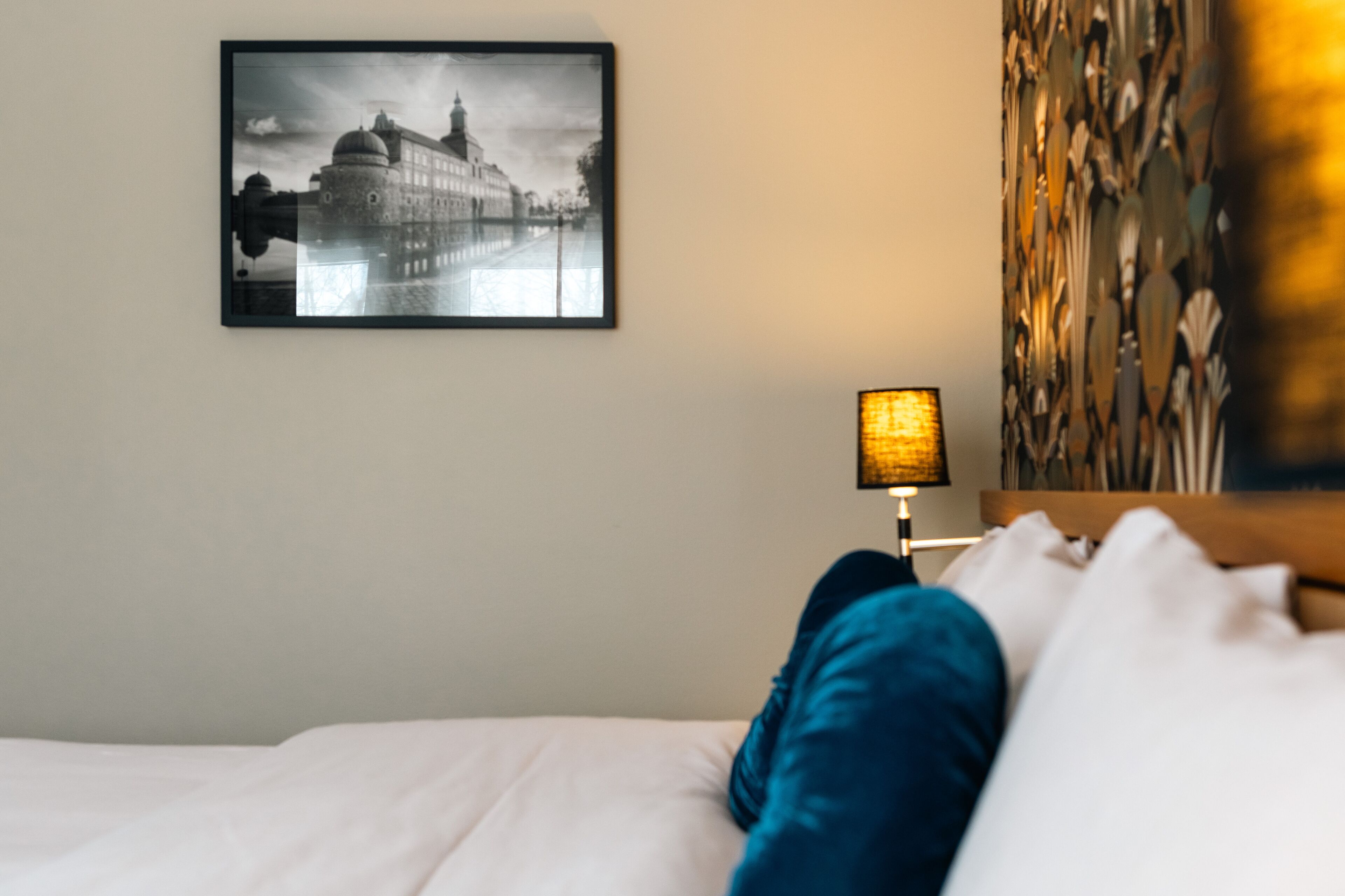 Ett hotellrum med en säng, en inramad tavla som föreställer en stadsbild, en lampa och en vägg med en intressant yta.