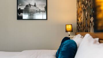 Ett hotellrum med en säng, en inramad tavla som föreställer en stadsbild, en lampa och en vägg med en intressant yta.