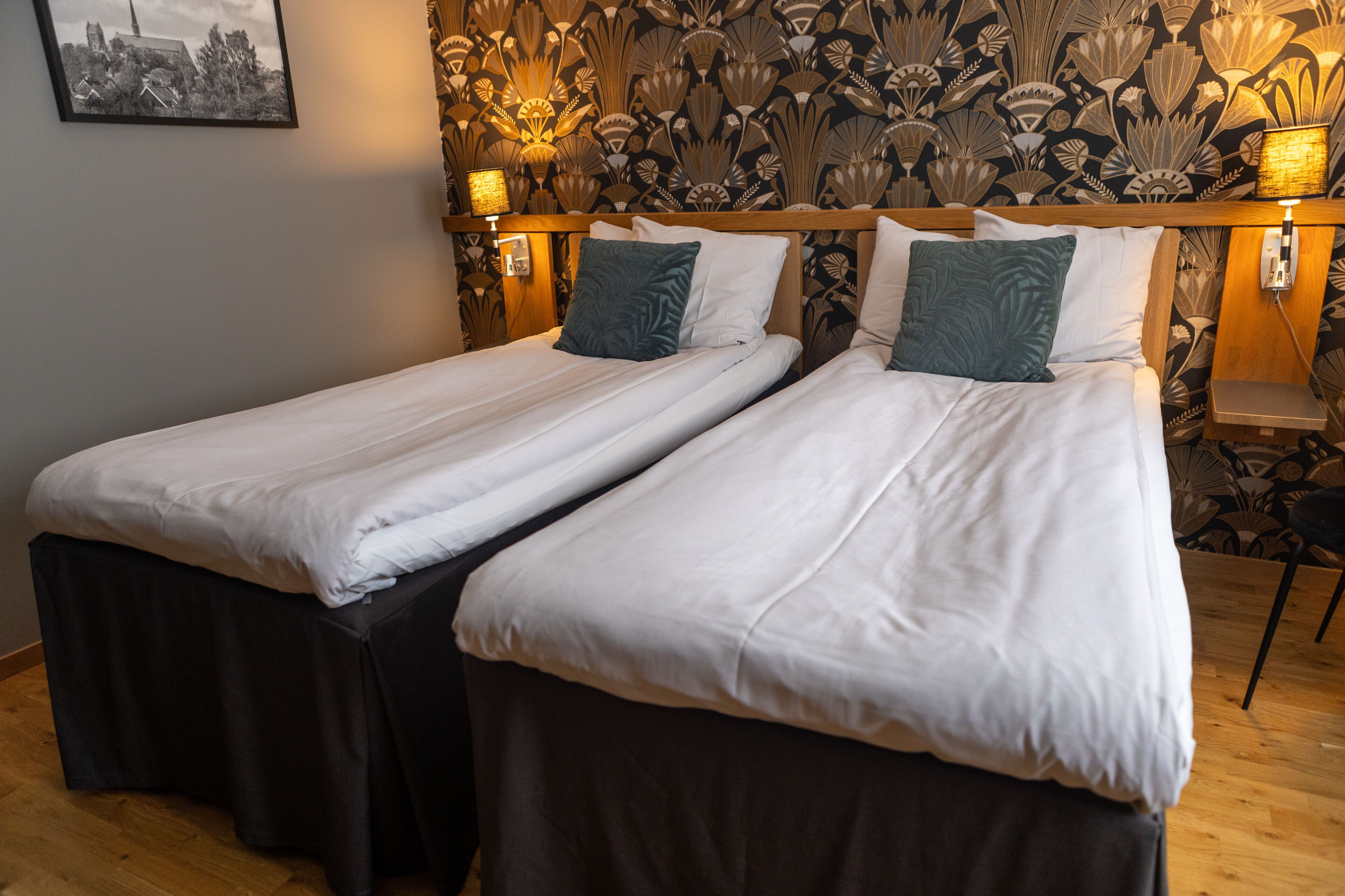 Un lit double avec du linge de lit blanc et une base de lit sombre dotée d’une jupe, deux oreillers et une lampe fixée au mur.