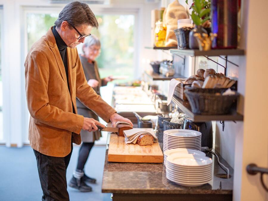 En person som skär bröd på en skärbräda i ett kök med ett fönster och en hylla med bröd och burkar.
