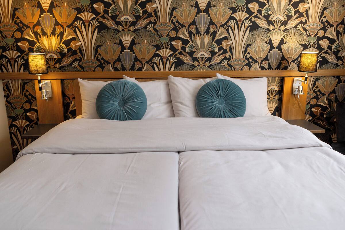 Ein Bett mit weißer Bettwäsche und zwei blauen Zierteilen, vor einer Wand mit floralem Wandpapier.