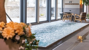 Un espace piscine intérieure avec vue sur l’extérieur, un coin salon avec des chaises et une plante en pot.