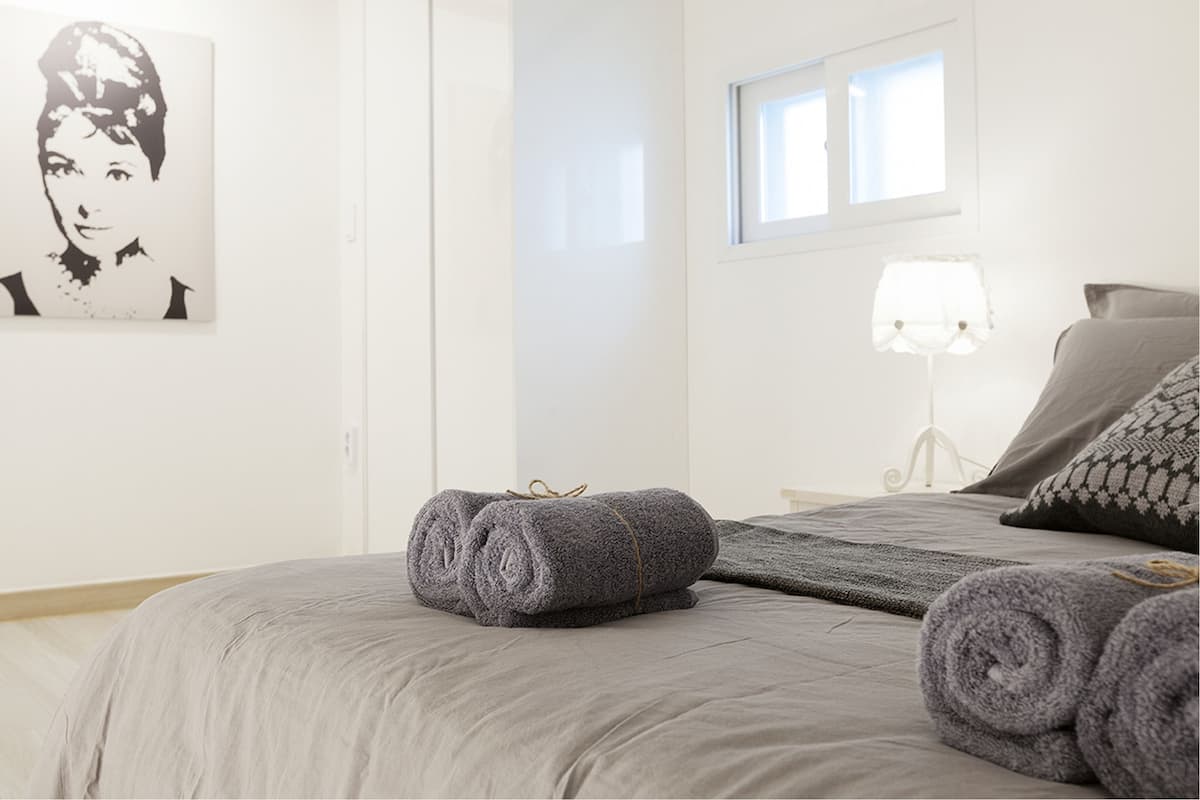 Ein ordentlich bezogenes Bett mit grauen Leinen und aufgerollten Handtüchern, eine Nachttischlampe und ein gerahmtes Kunstwerk an der Wand.