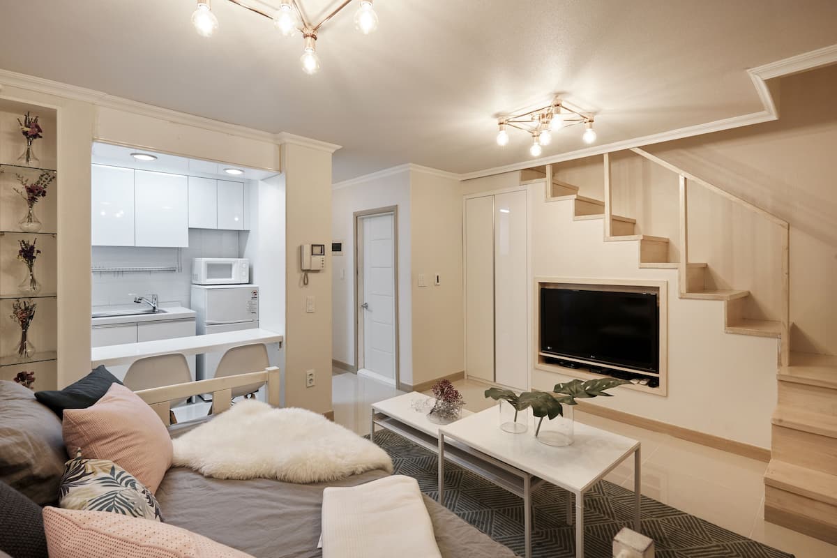 Ein modernes Wohnzimmer mit einer Couch, einem Couchtisch und einem an der Wand befestigten Fernseher. Eine Treppe führt zu einer oberen Etage, und durch eine offene Tür ist ein Küchenbereich sichtbar.