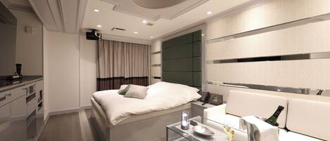 A modern hotel room with a bed, a glass table with a bottle and glasses, a sofa, and a minibar.