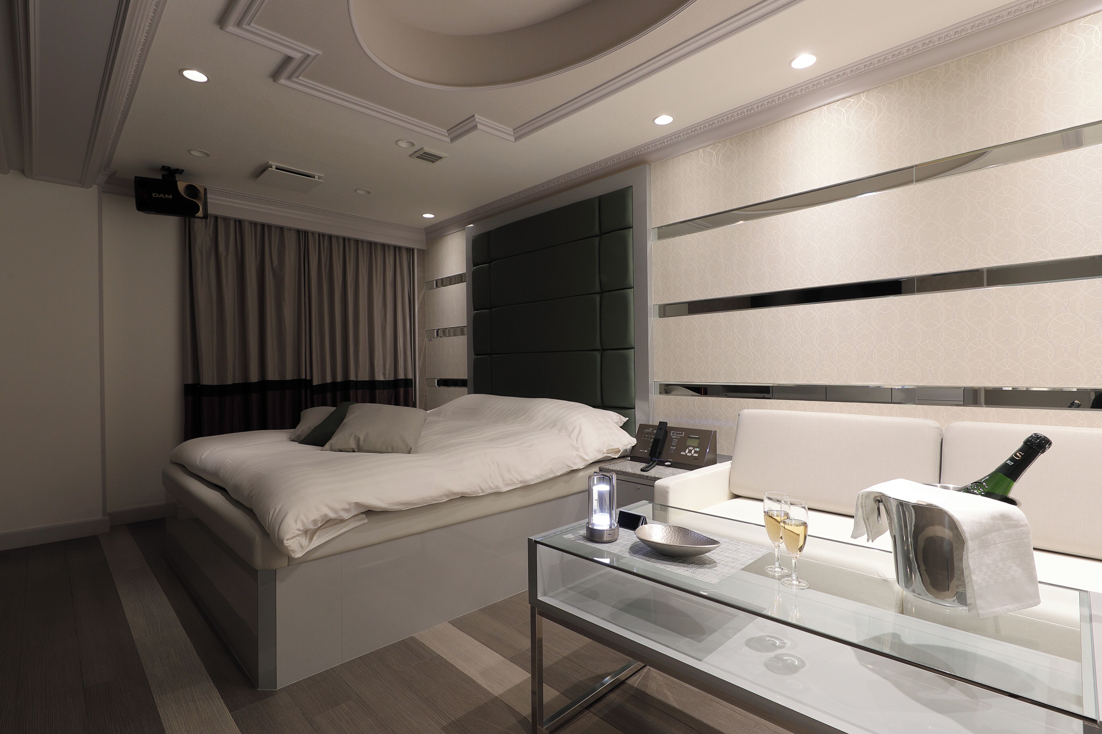 A modern hotel room with a bed, a glass table with a bottle of champagne, a bedside table with a lamp, and a wall-mounted TV.