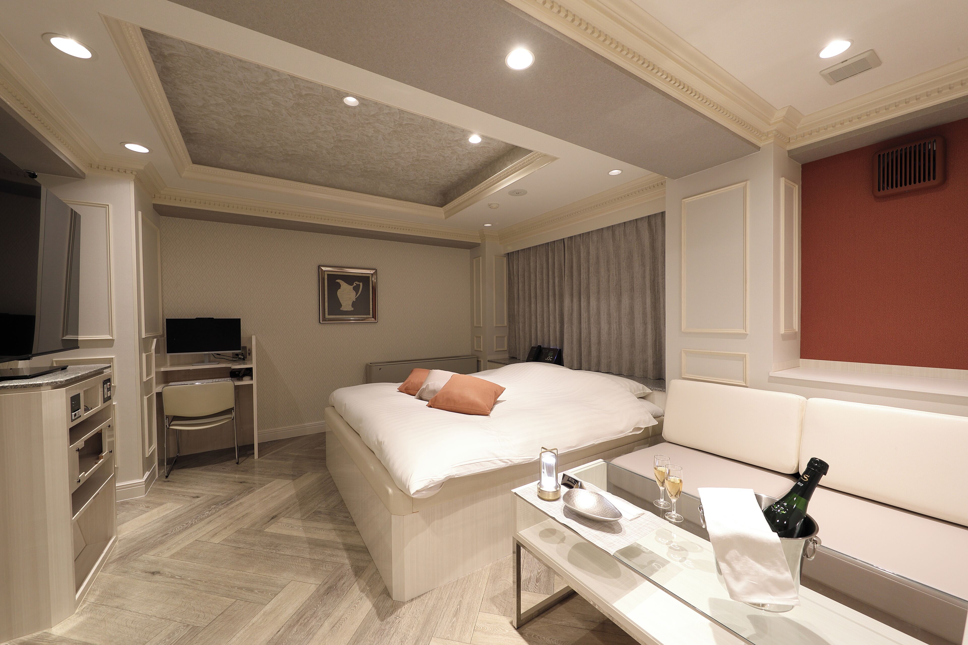A hotel room with a bed, desk, chair, and a minibar with a bottle and glasses.