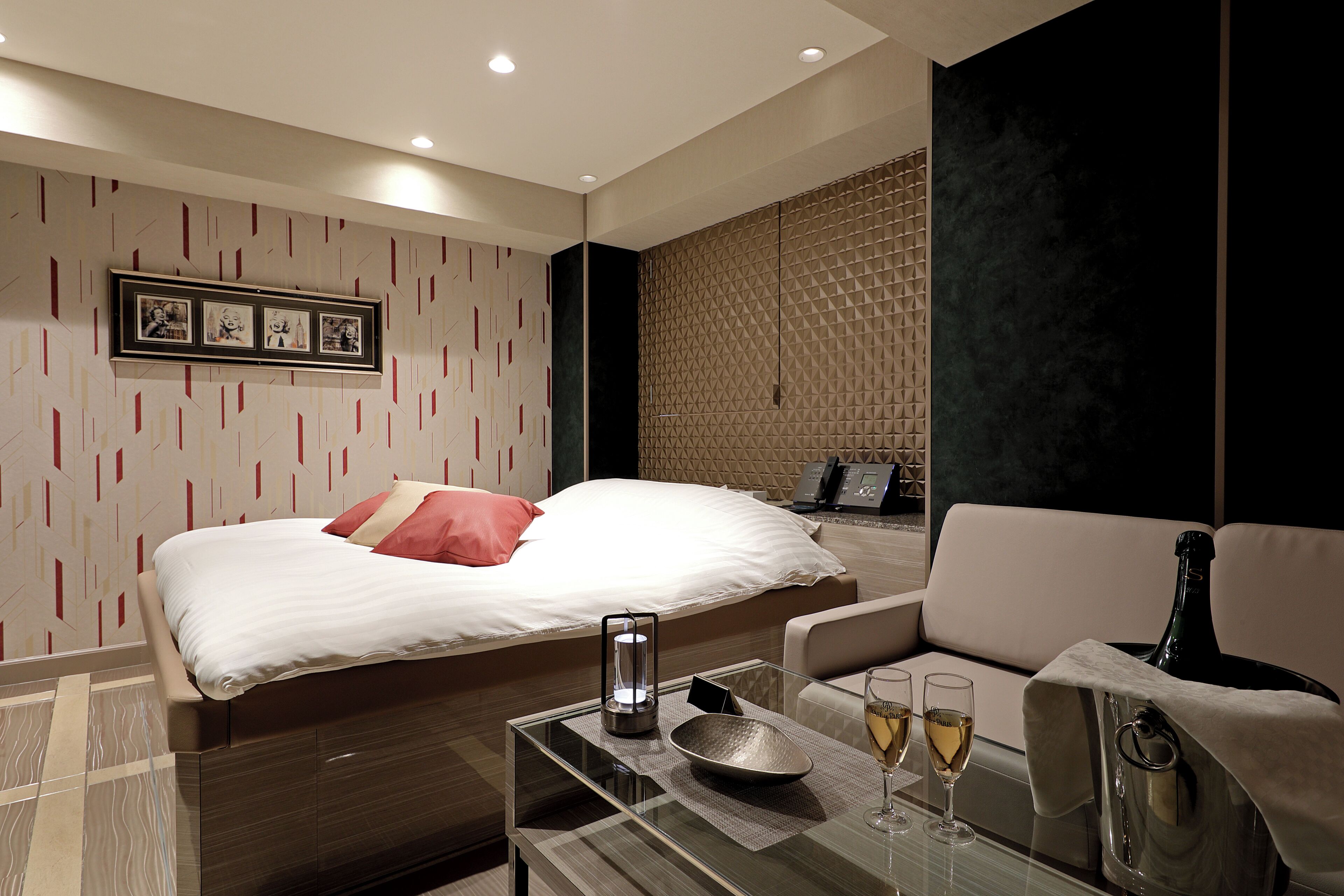A modern hotel room with a neatly made bed, a glass coffee table with a bottle and glasses, and a seating area with a couch and armchair.