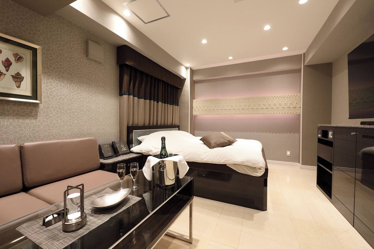 Una habitación de hotel moderna con cama, sofá, minibar y una mesita con una botella y un vaso.