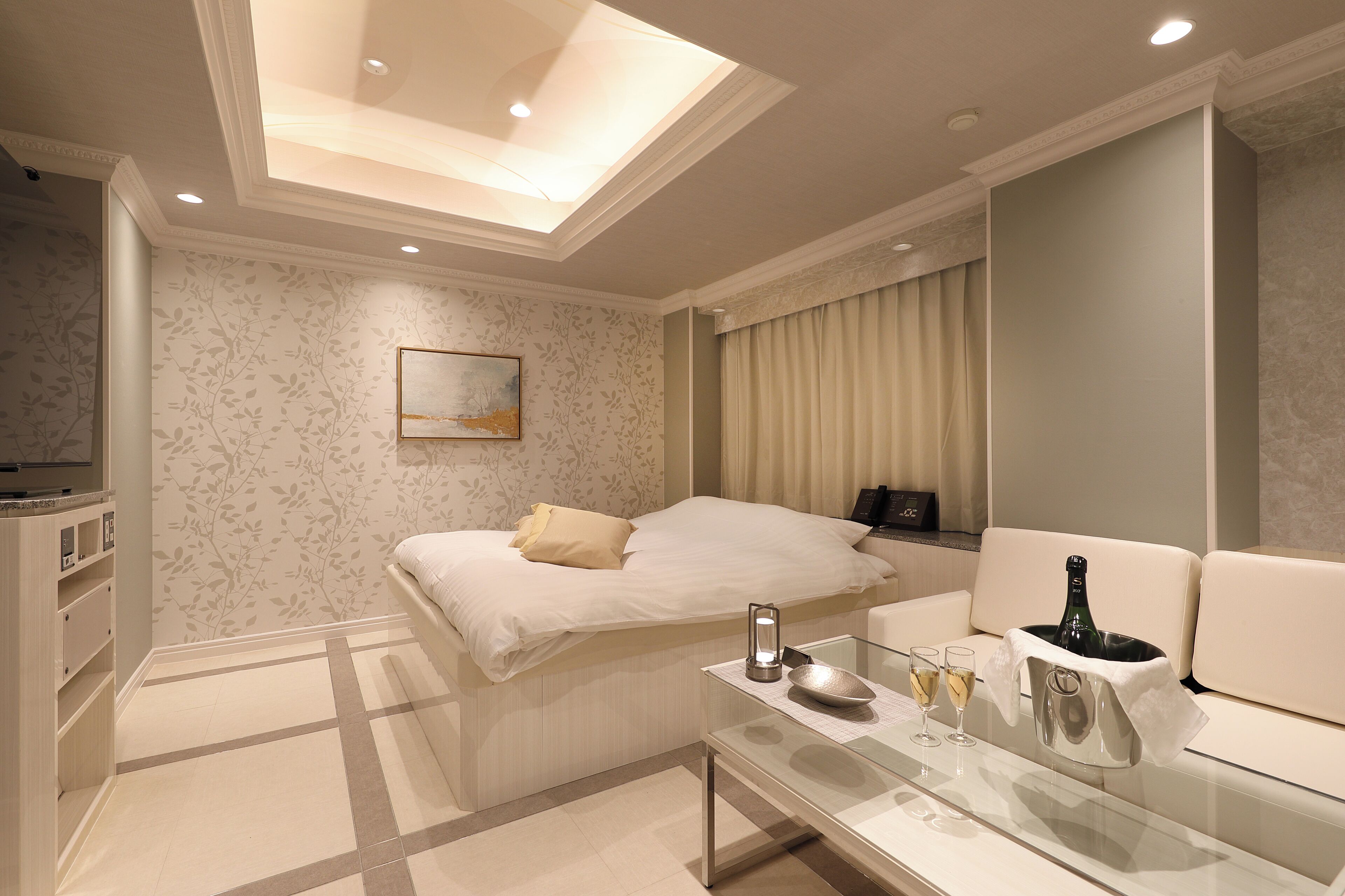 A modern hotel room with a bed, a glass coffee table with a bottle and glasses, a painting on the wall, and a ceiling light.